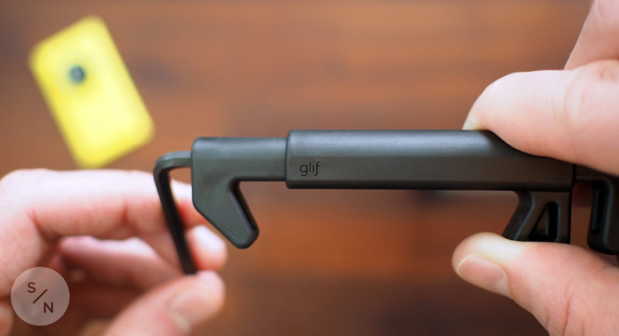 A person holding an adjustable GLIF tripod mount with a blurred yellow smartphone in a case in the background.
