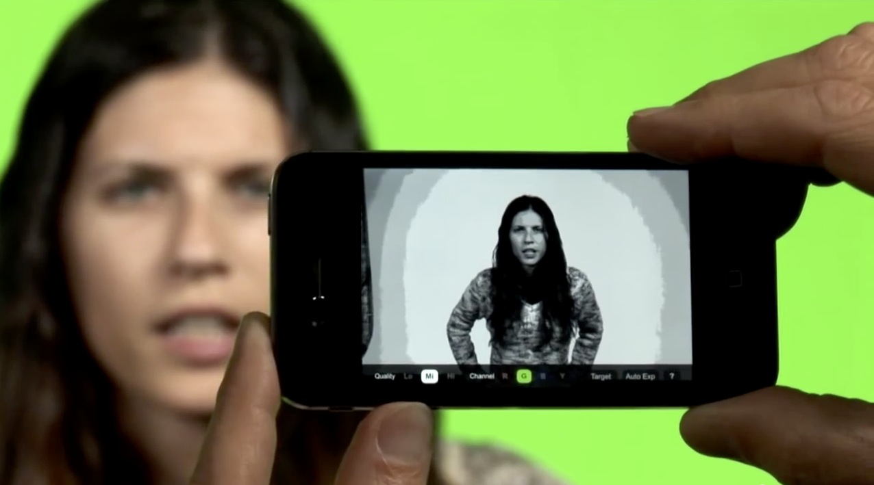 A person holds a smartphone displaying a video of a woman, with the real woman in the background expressing concern or confusion against a green screen.