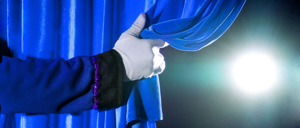 A hand in a white glove pulling back a blue stage curtain with a bright light in the background, heralding a revolution.