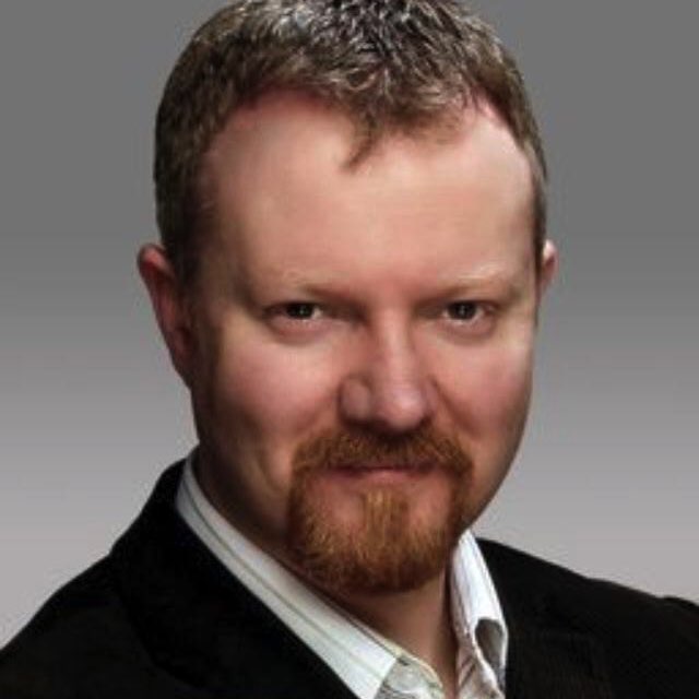 A portrait of a man with a beard and mustache, wearing a black jacket over a white shirt.