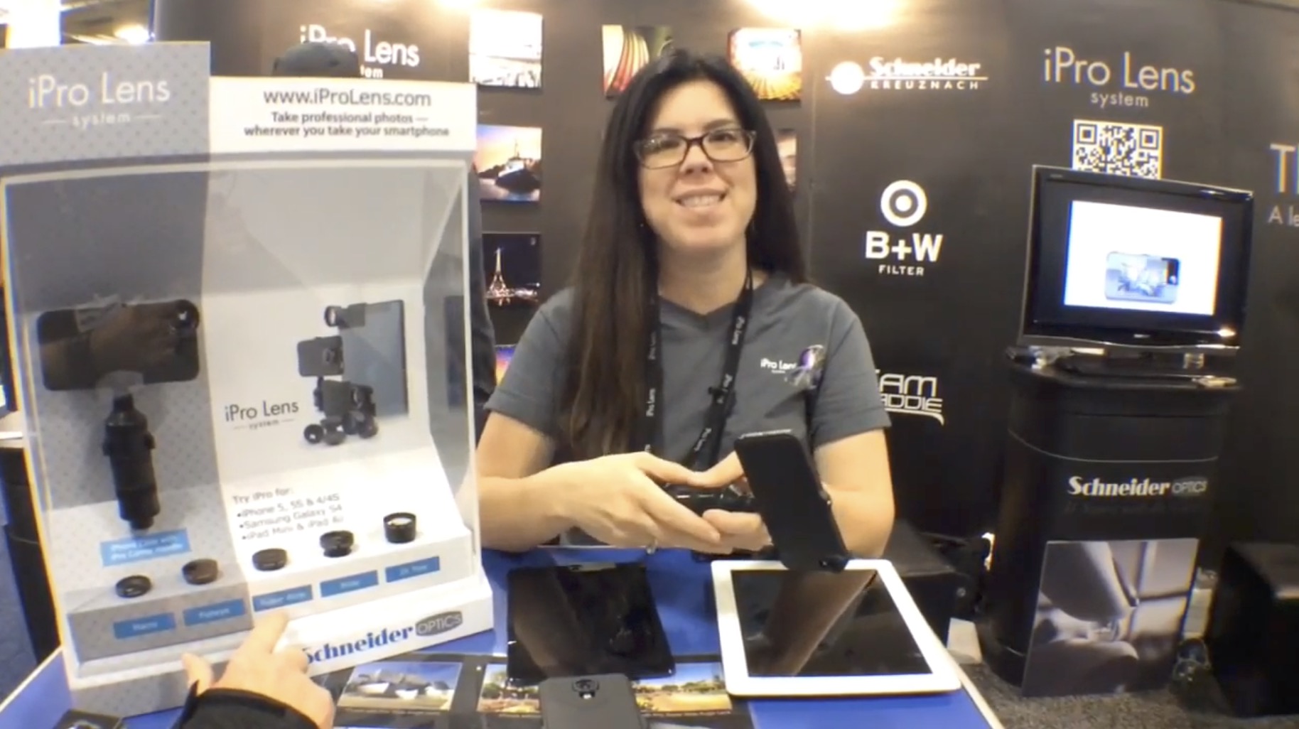 A smiling woman demonstrating a Samsung mobile photography lens at a trade booth.