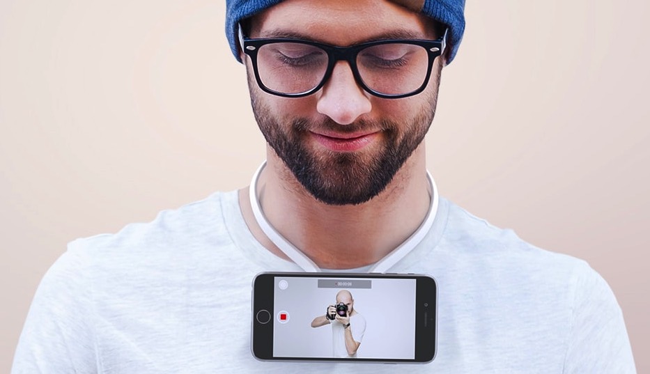 Man in glasses and beanie gearing up, smiling down at smartphone camera ready to start live video streaming.