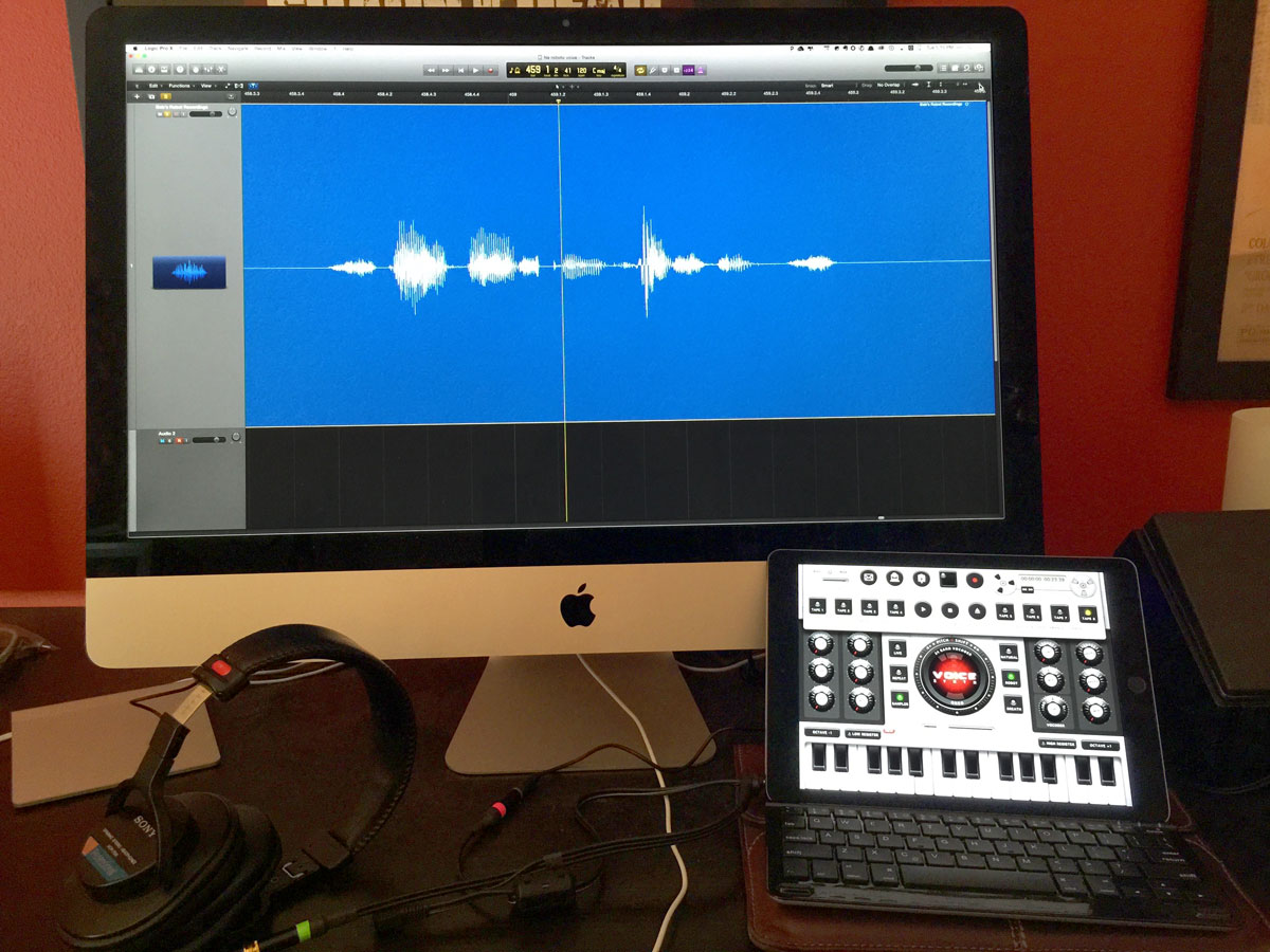 A desktop workstation with audio editing software open on an iMac, accompanied by a MIDI controller, headphones, and a voice synth.